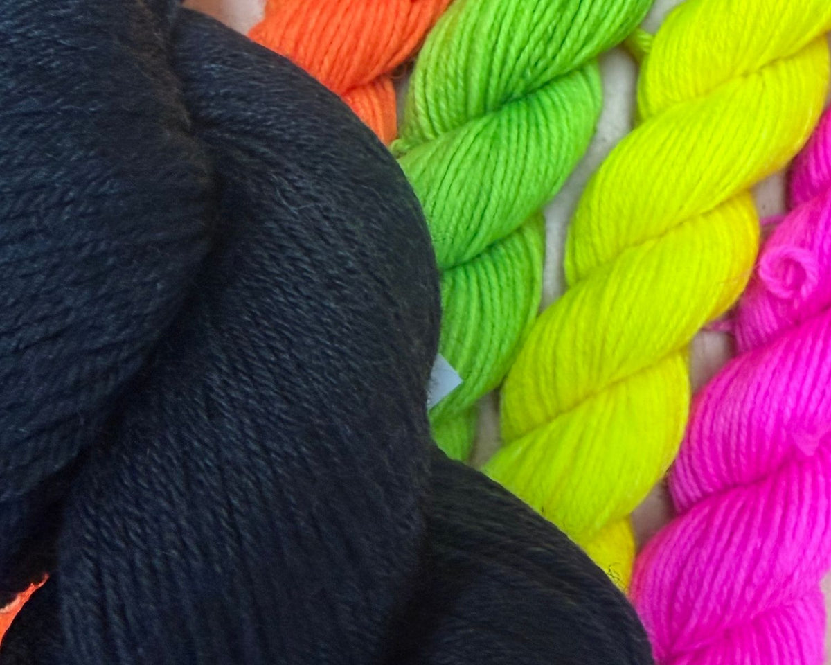 Close-up of twisted yarn skeins in multiple bright colors, including black in the foreground, orange, neon green, neon yellow, and hot pink.
